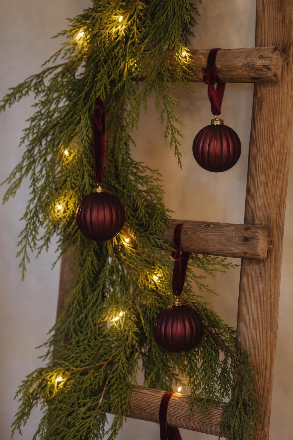 Close view of a cedar garland woven diagonally through ladder rungs with warm white lights and three matte burgundy ornaments hanging on ribbon