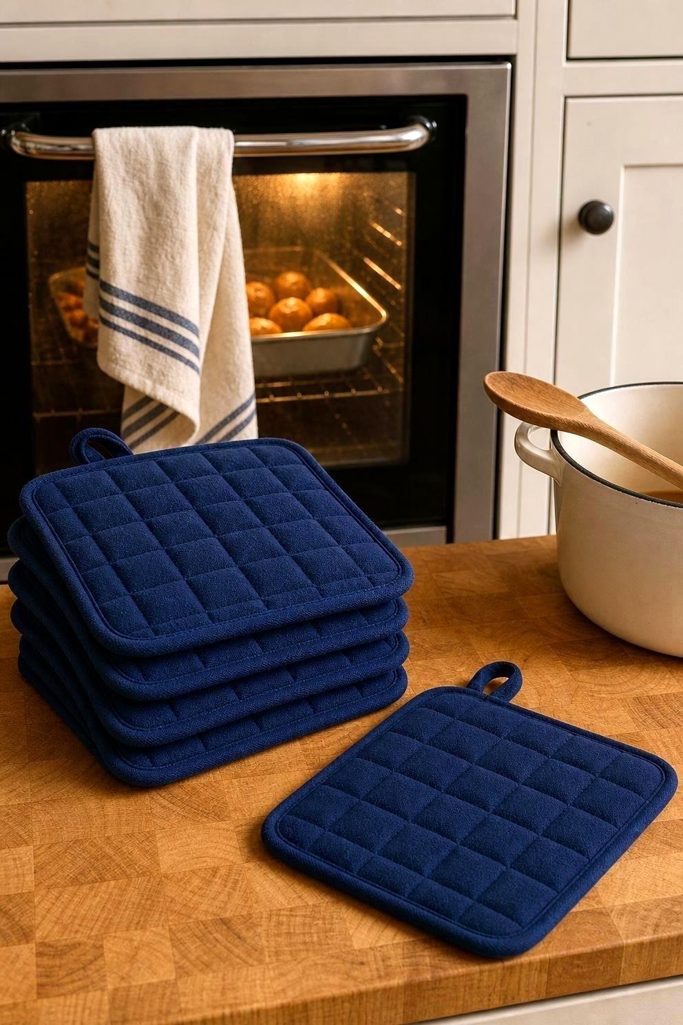 Five folded cotton pot holders in dark blue stacked on a wooden kitchen counter next to a warm oven with a roasting tin visible, a cotton towel over the oven handle