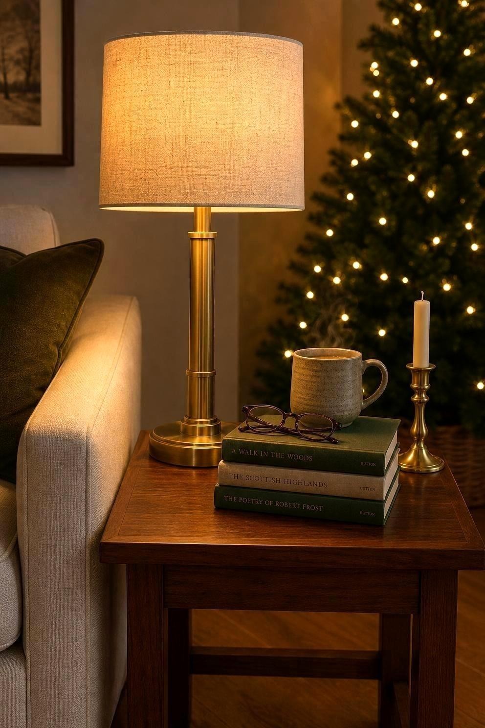 A brass table lamp with a linen shade lit beside a stack of books and a small brass candle holder, a lit Christmas tree softly out of focus behind