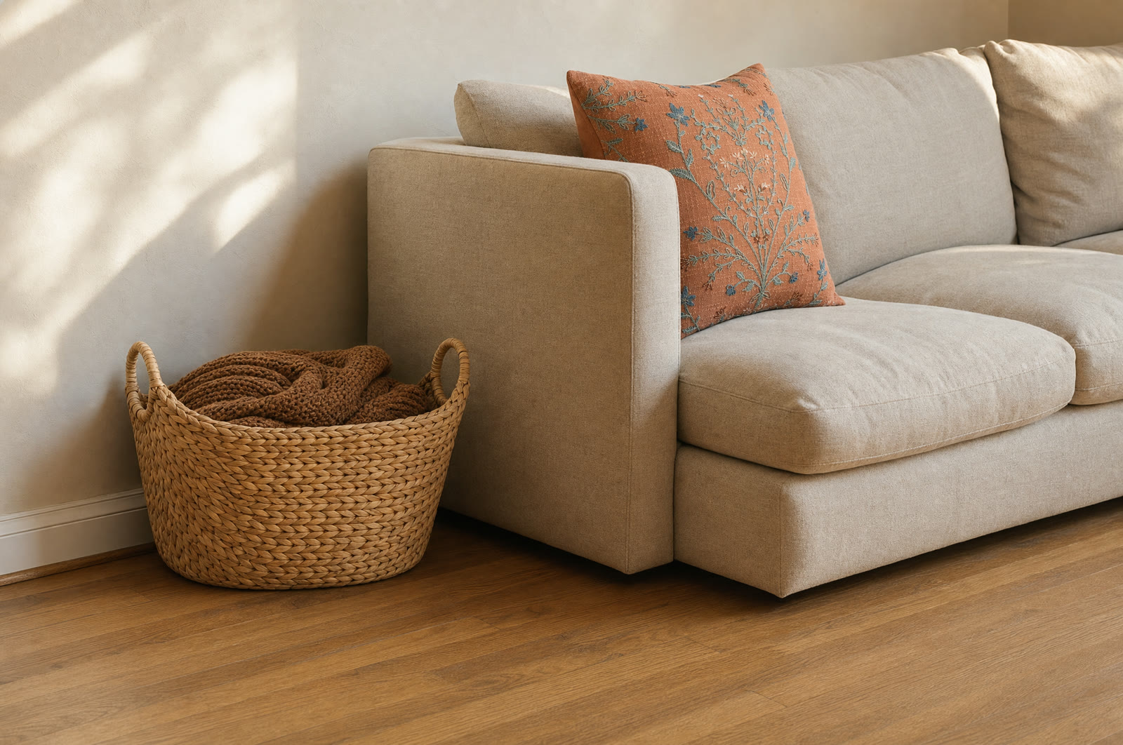 Natural woven water-hyacinth basket holding a brown knit throw beside the arm of a cream sofa with a terracotta floral pillow