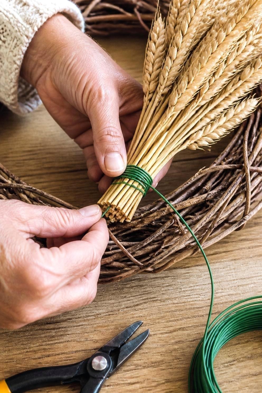 Close view of hands wrapping green floral wire around the cut stem ends of a small wheat bunch against a grapevine wreath base