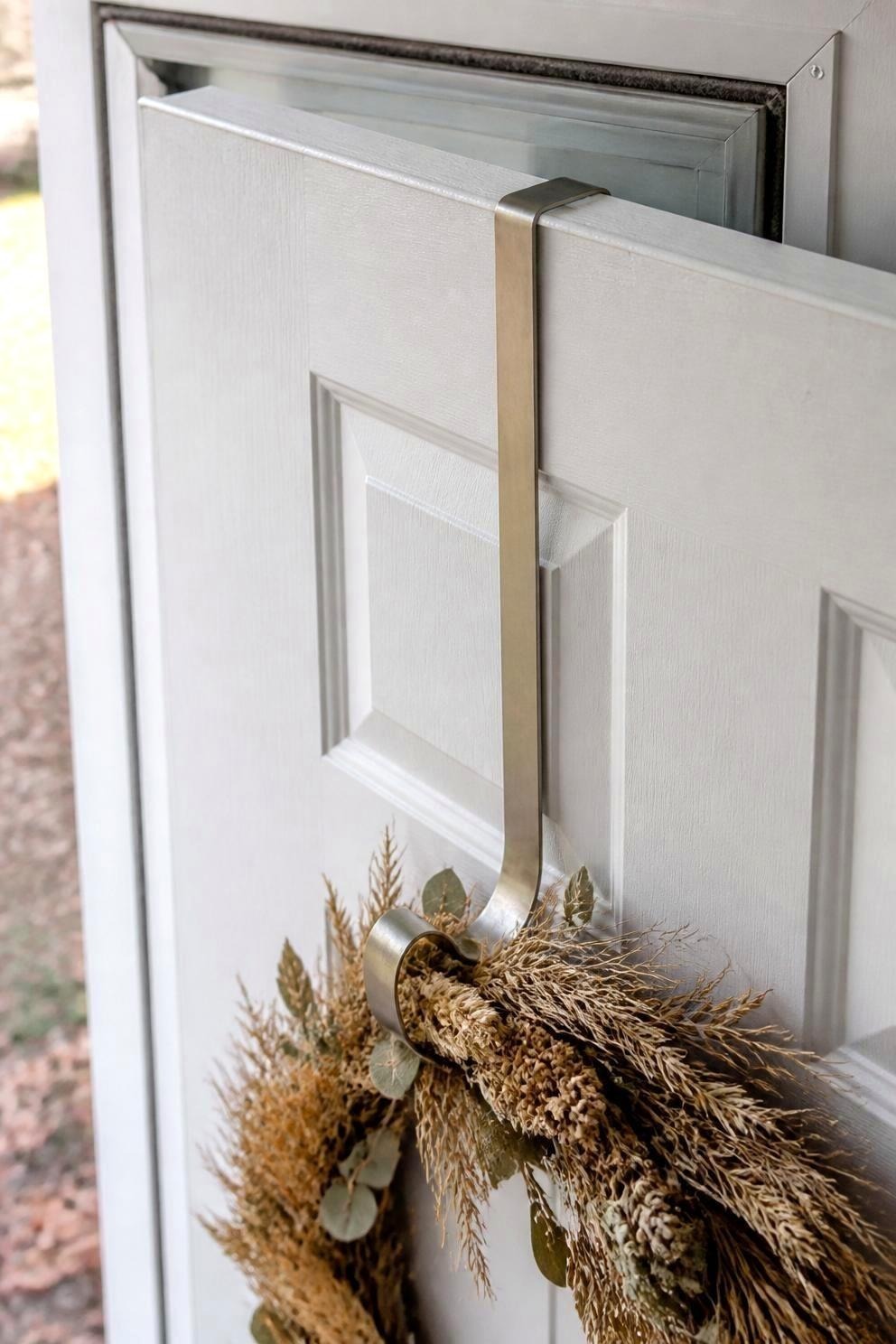 A slim over-the-door wreath hook seen from above at the top edge of a white door, holding a neutral dried wreath, with a storm door closed just beyond it