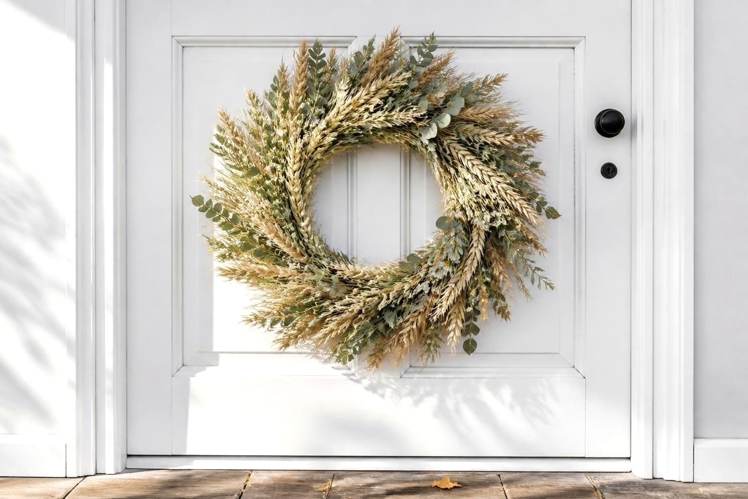 A dried wheat and eucalyptus wreath in cream and sage hanging on a white front door, no orange, cool afternoon daylight from the left