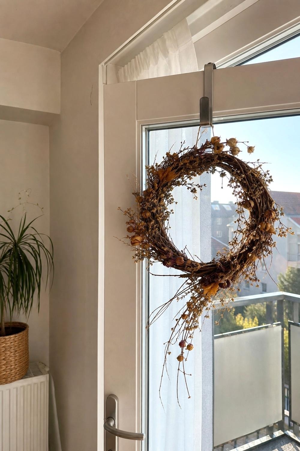 Close-up of an over-door hook holding a lightweight fall wreath on a balcony door, with a fiberglass planter of ornamental kale nearby