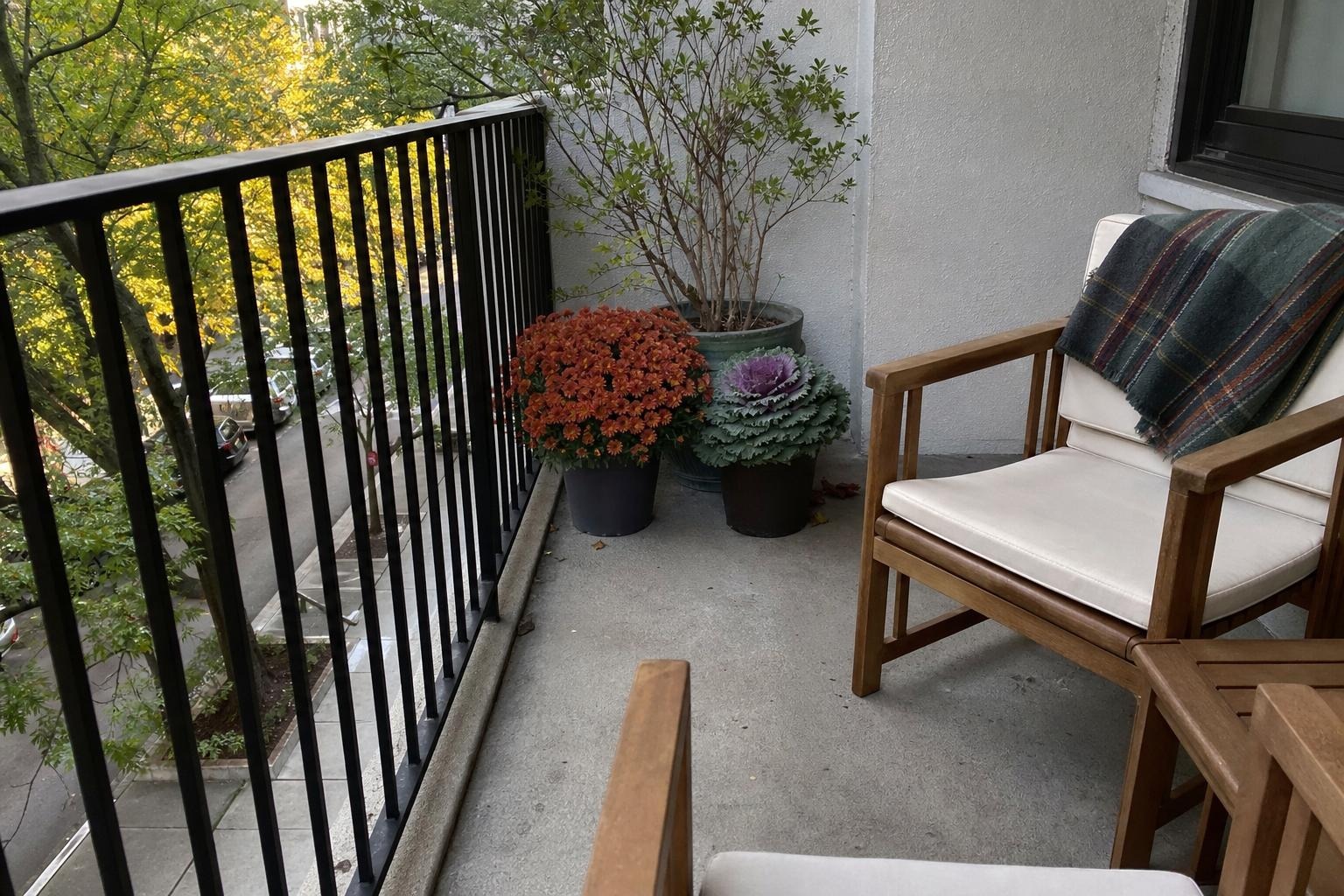 Small apartment balcony in fall with potted mums and ornamental kale, a folded wool throw on a chair, and a small side table