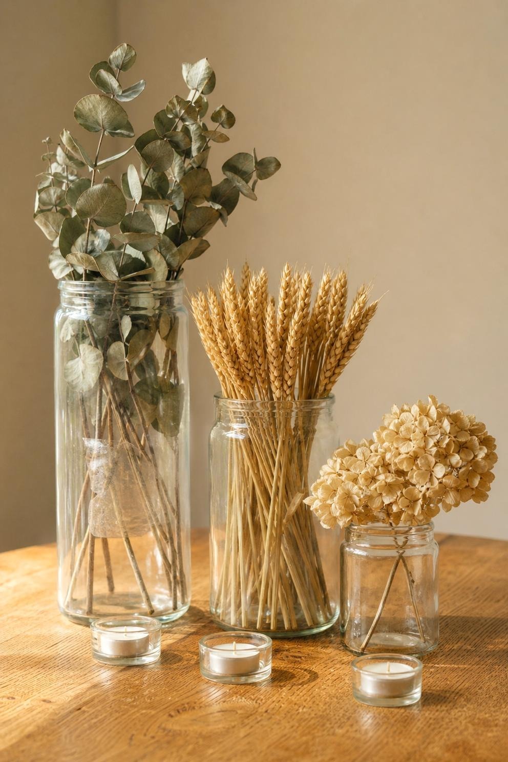 Three plain glass jars of different heights holding dried eucalyptus and wheat, grouped on a wooden table with tealights