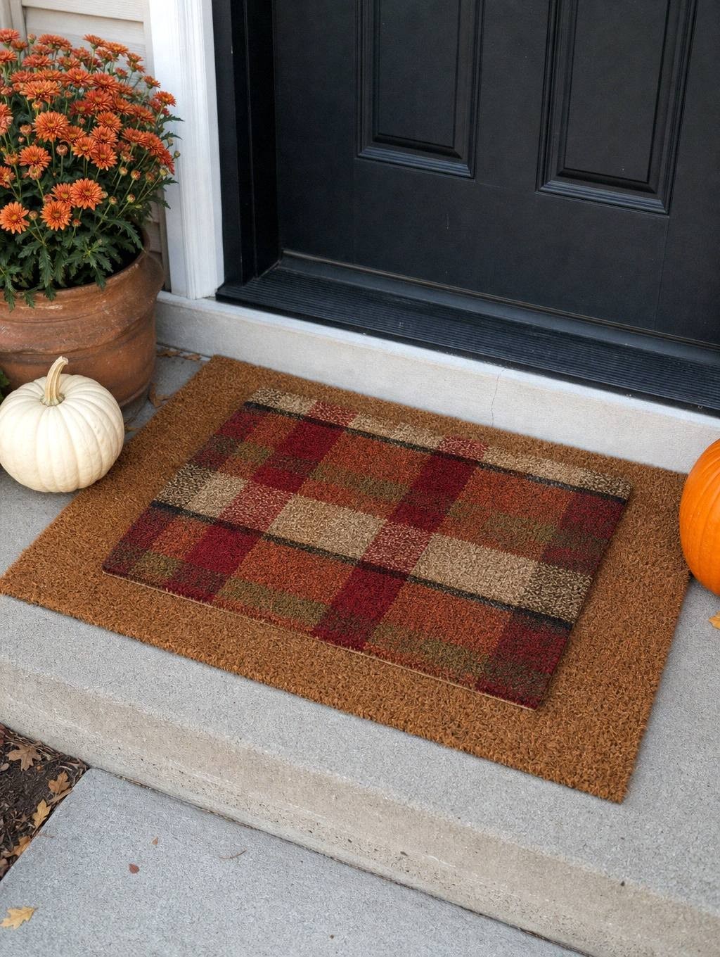 coir doormat layered under a smaller plaid mat on a small front step beside orange mums and a white pumpkin