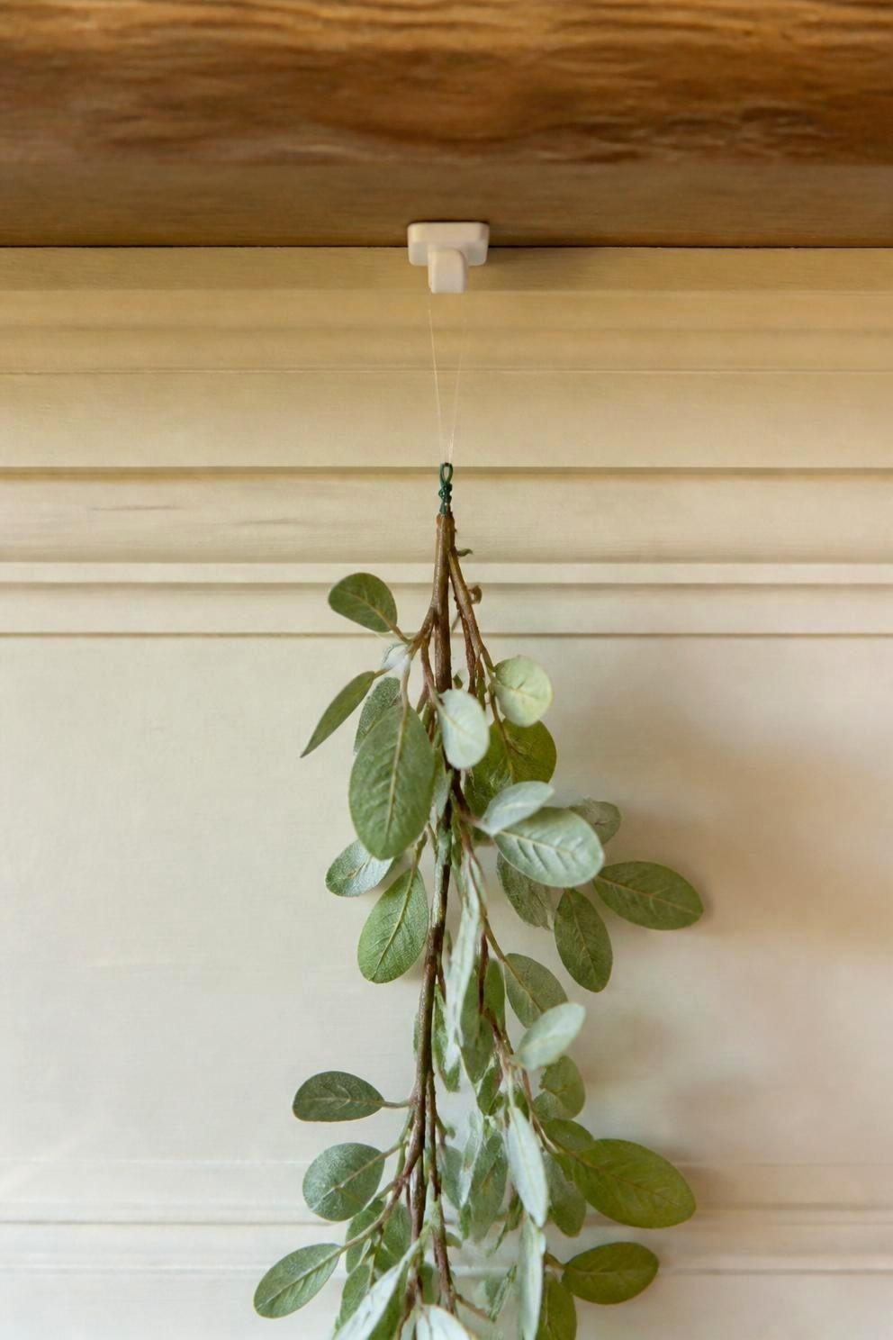 Close view of a small white adhesive hook pressed to the underside of a wooden mantel shelf with a length of clear line looped over it and a garland stem wired to the line