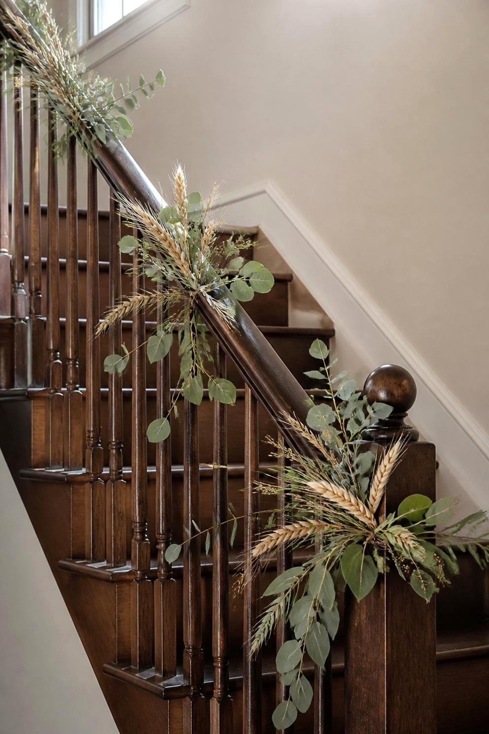 A muted green and dried wheat garland spiraled loosely around a dark wooden stair banister with wraps about a foot apart and a fuller cluster gathered at the newel post
