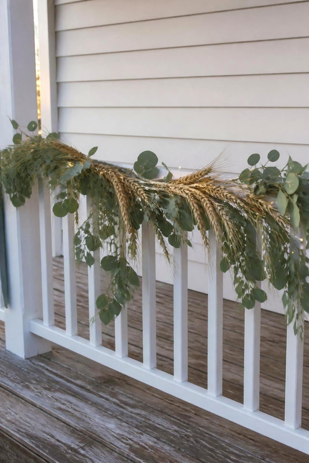 A eucalyptus and wheat garland wired along a white porch rail with a warm white fairy light strand woven through it, glowing at dusk against weathered porch boards