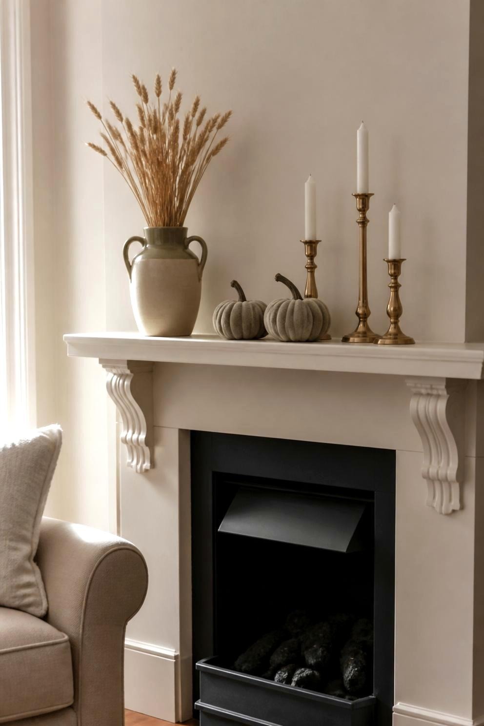 A neutral mantel in cream, tan, and brass with trimmed wheat in a handled ceramic vase, three brass candlesticks of different heights, and two pale gray pumpkins, no orange anywhere