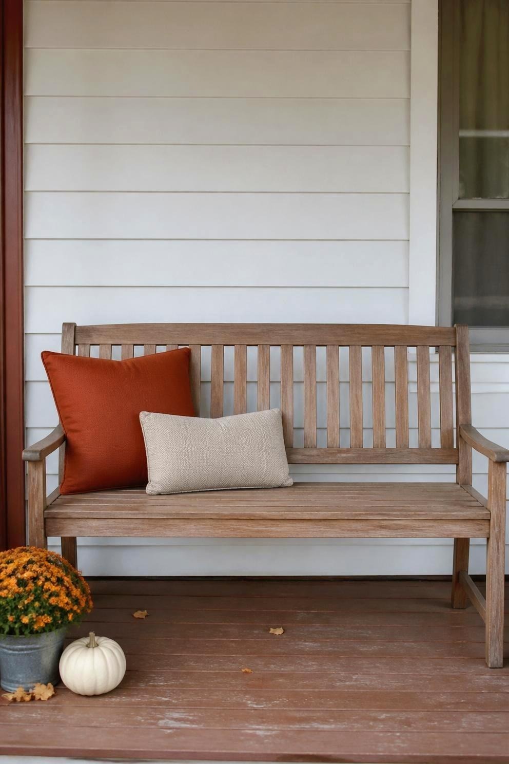 A four foot wooden porch bench holding one large rust square pillow and one small oatmeal lumbar pillow, with an empty seat beside them