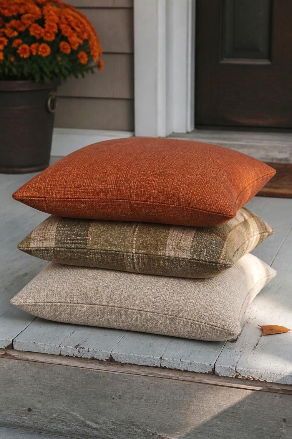 Three outdoor pillows in rust, muted olive plaid, and warm oatmeal stacked on a wooden porch step in cool afternoon light