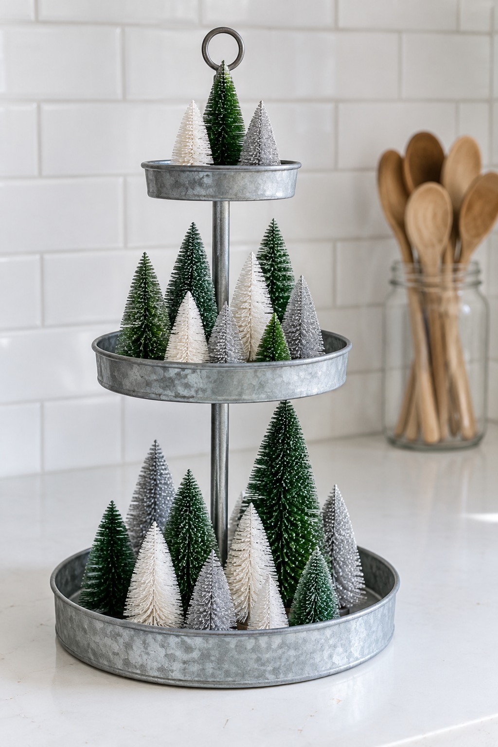 Galvanized three tier stand on a white kitchen counter holding green, white and silver bottle-brush trees of different heights, with a white tile backsplash and a wooden spoon jar behind