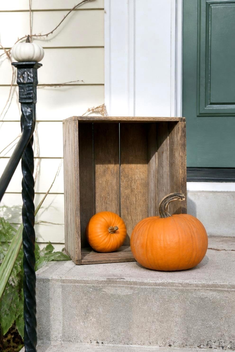 A narrow apartment stoop with a wooden crate turned upside down holding a small pumpkin, a medium pumpkin on the concrete at its base, and a miniature white pumpkin on the railing post