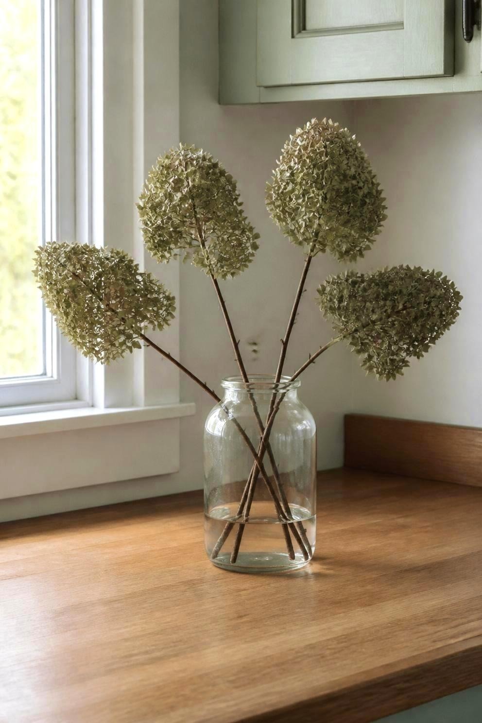 Four leafless hydrangea stems standing in a clear glass jar with two inches of water at the bottom, set on a kitchen counter away from the window