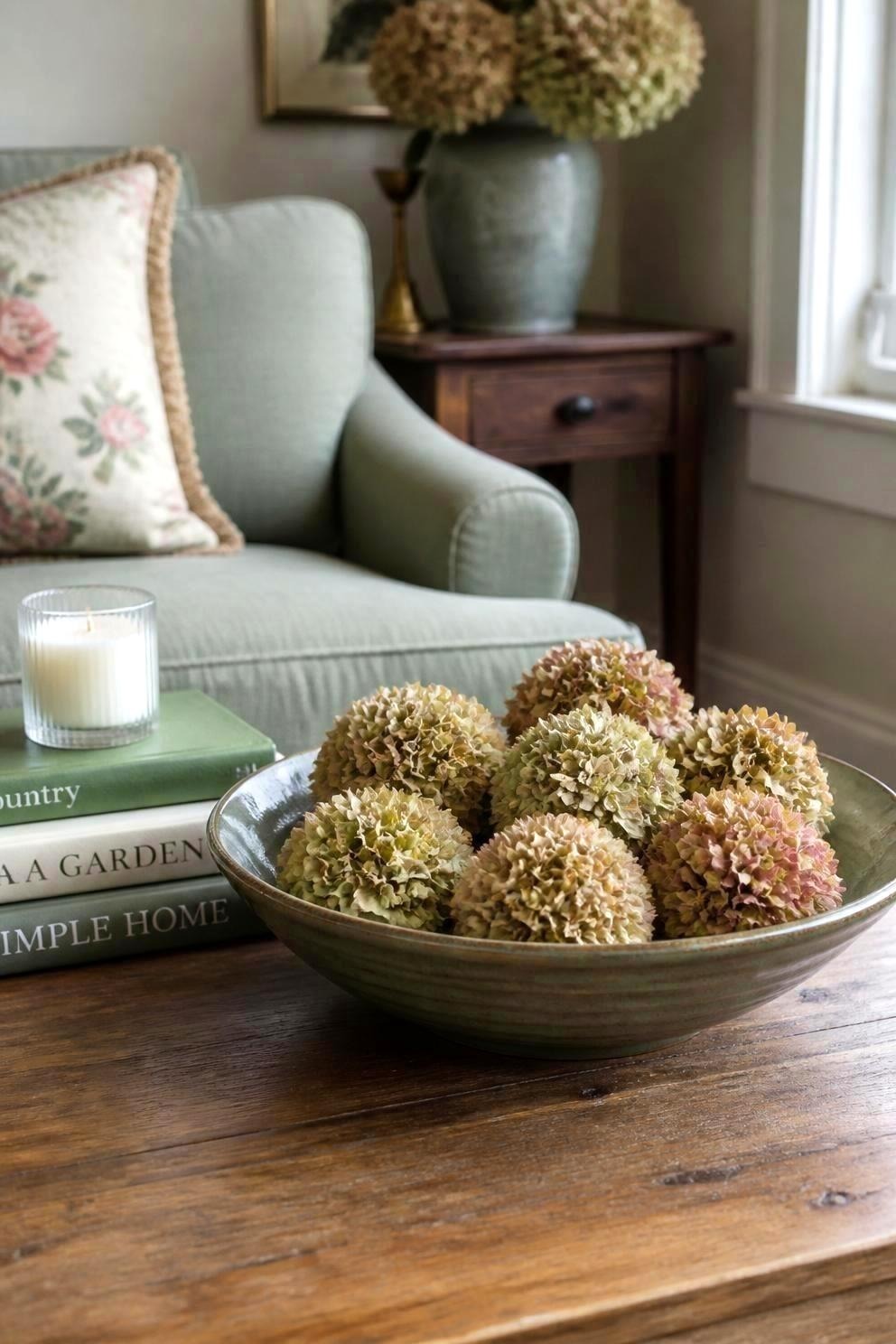 Seven dried hydrangea heads with short stems arranged in a wide shallow ceramic bowl on a coffee table beside a stack of books and a lit candle