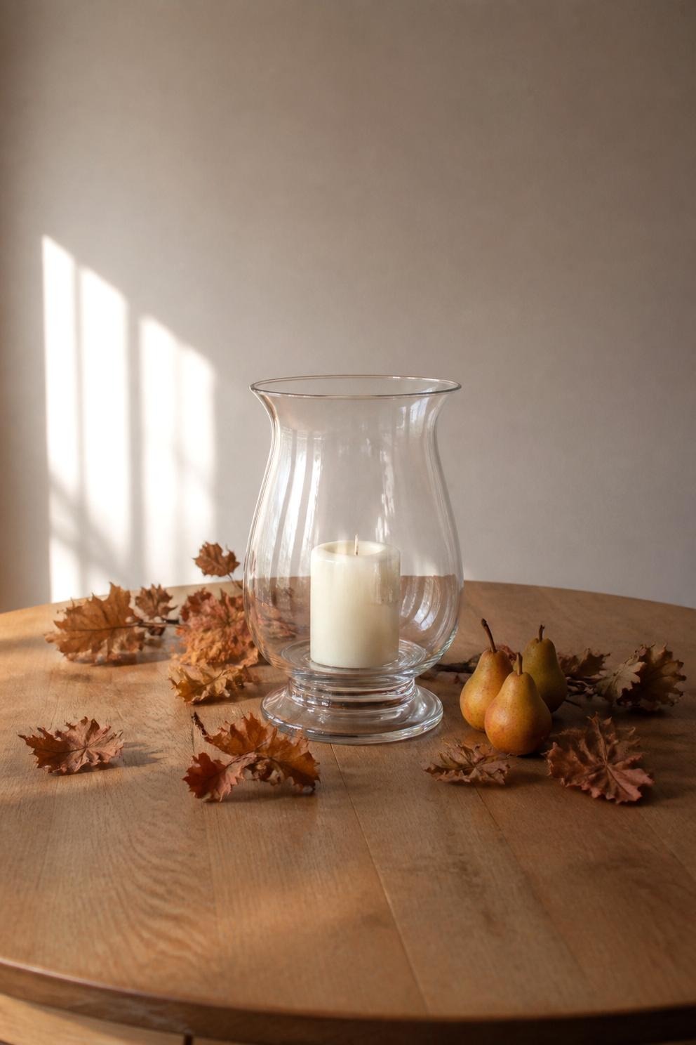 A single glass hurricane candleholder at the centre of a dining table with a low line of dried foliage and two small pears