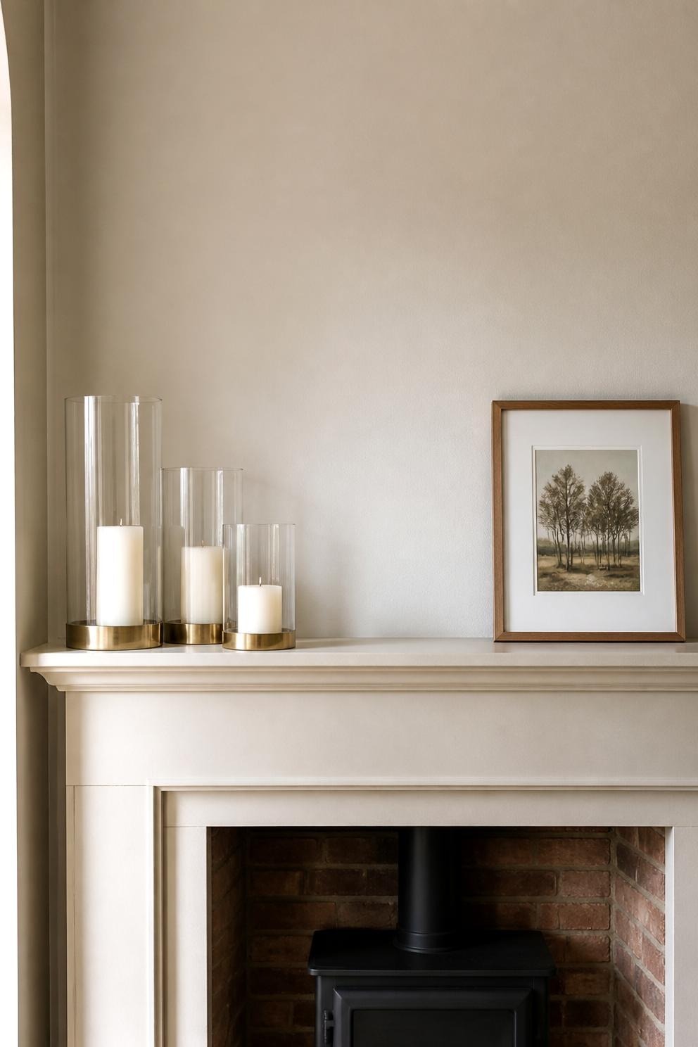 Three hurricane candleholders clustered at one end of a mantel, balanced by a framed print at the other end