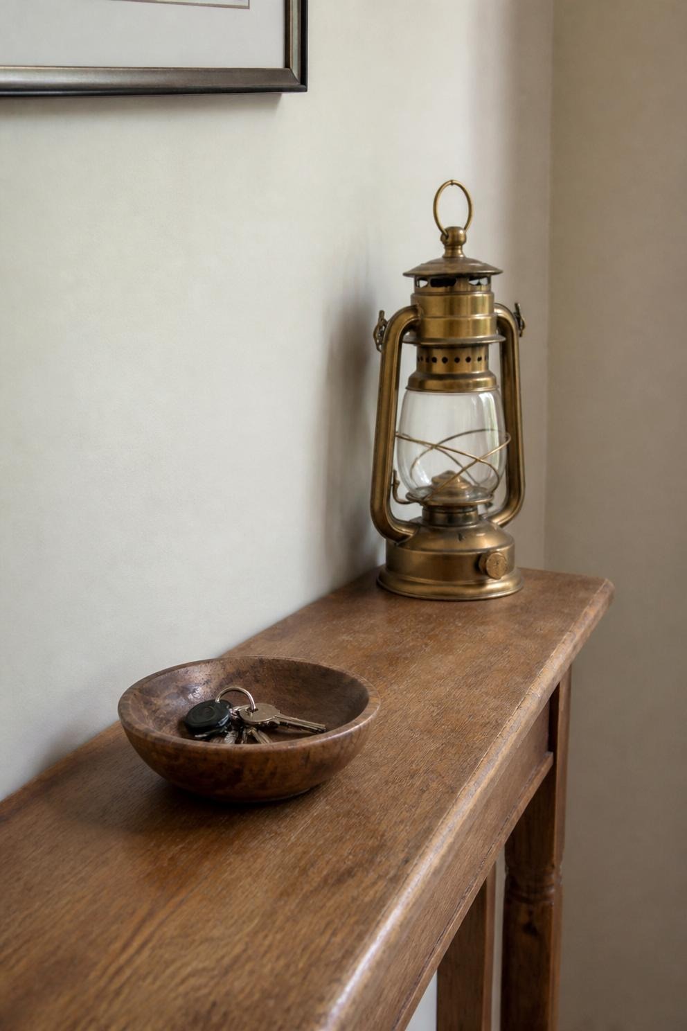 A compact hurricane lantern at the back corner of an entry console beside a shallow wooden bowl holding keys