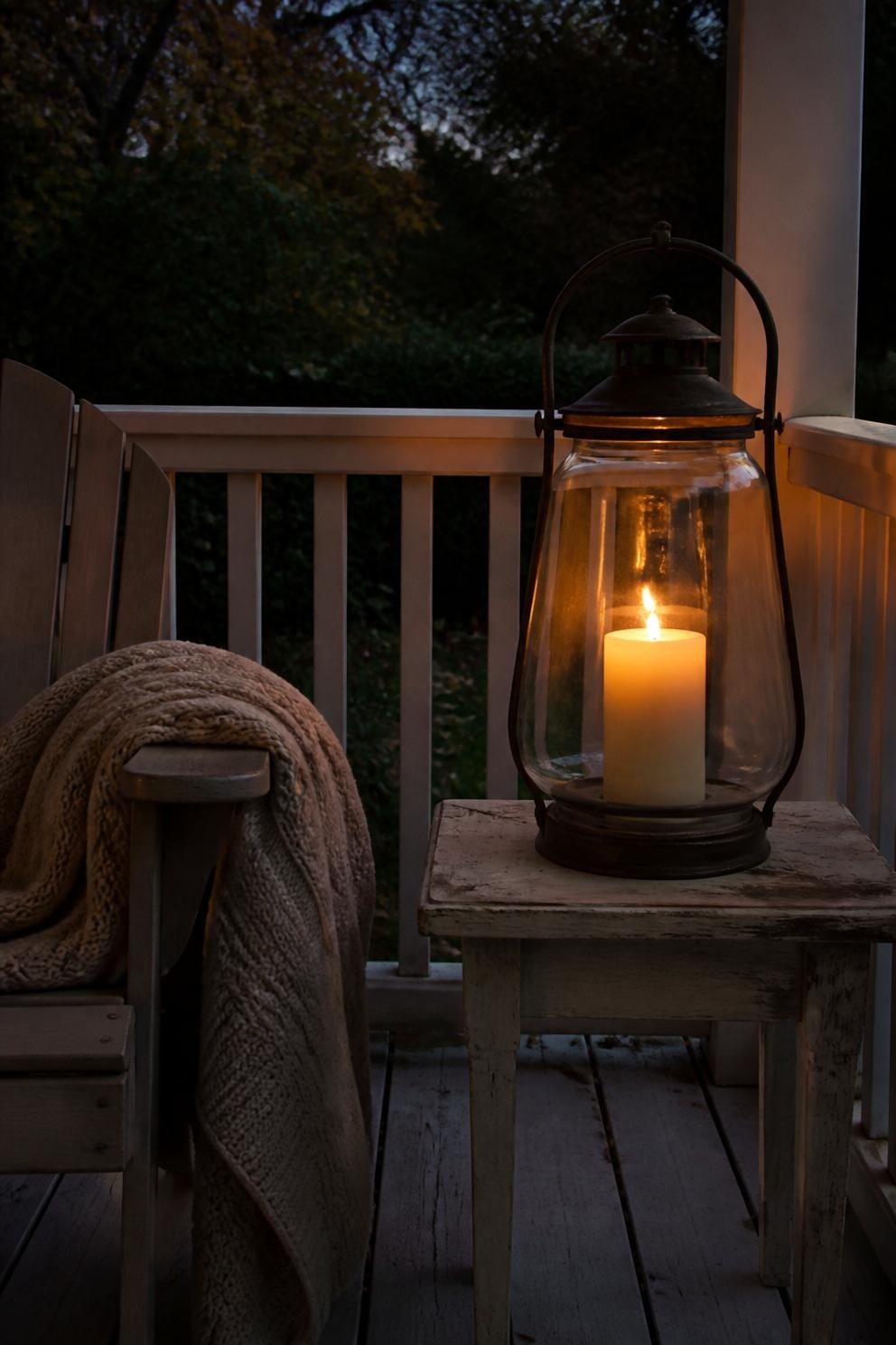 A hurricane lantern with a flameless candle on a sheltered porch table at dusk beside a folded wool throw