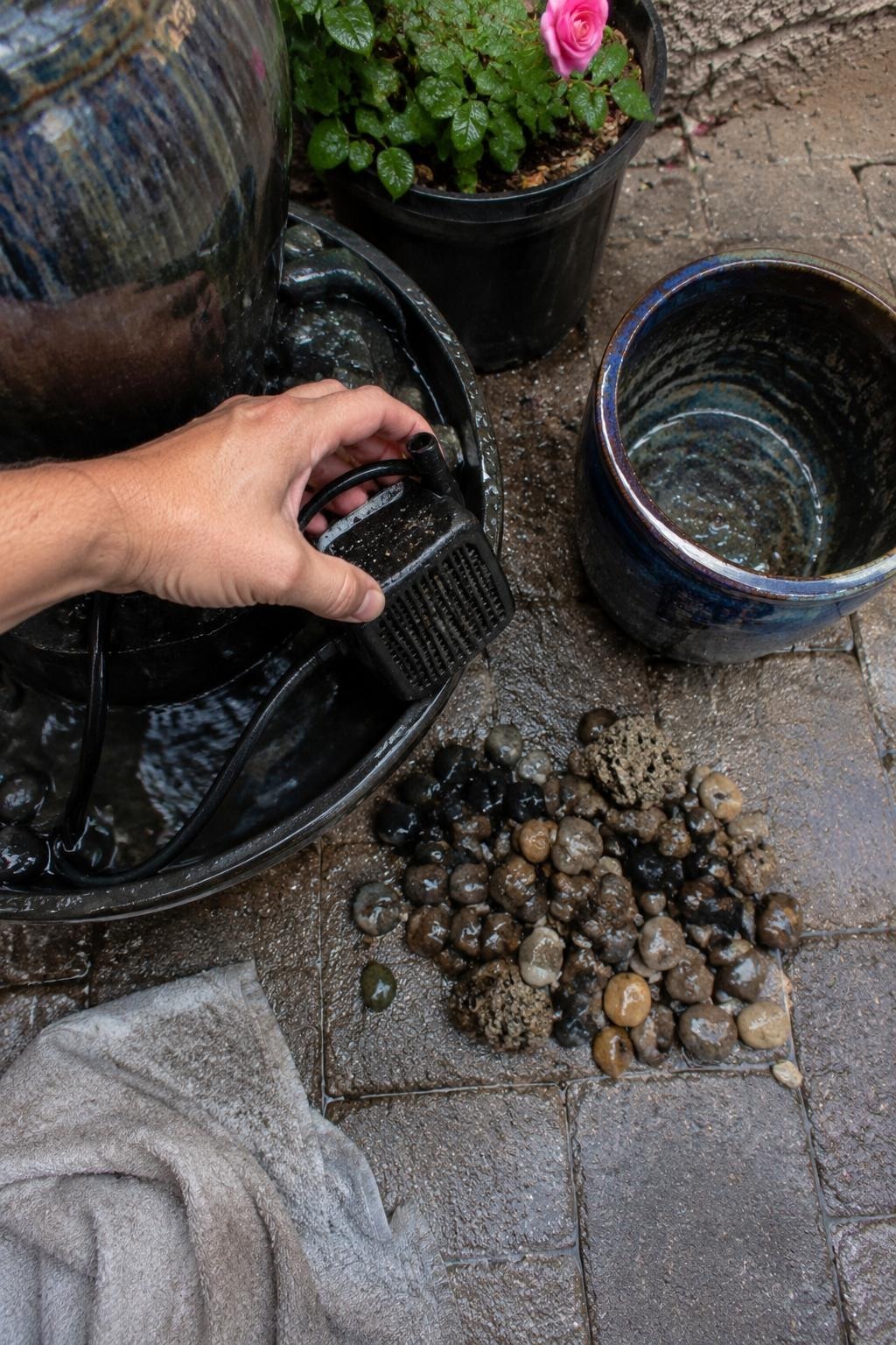 A hand lifting a small fountain pump out of a reservoir basin to rinse the intake screen, gravel and a glazed pot beside it on a paver
