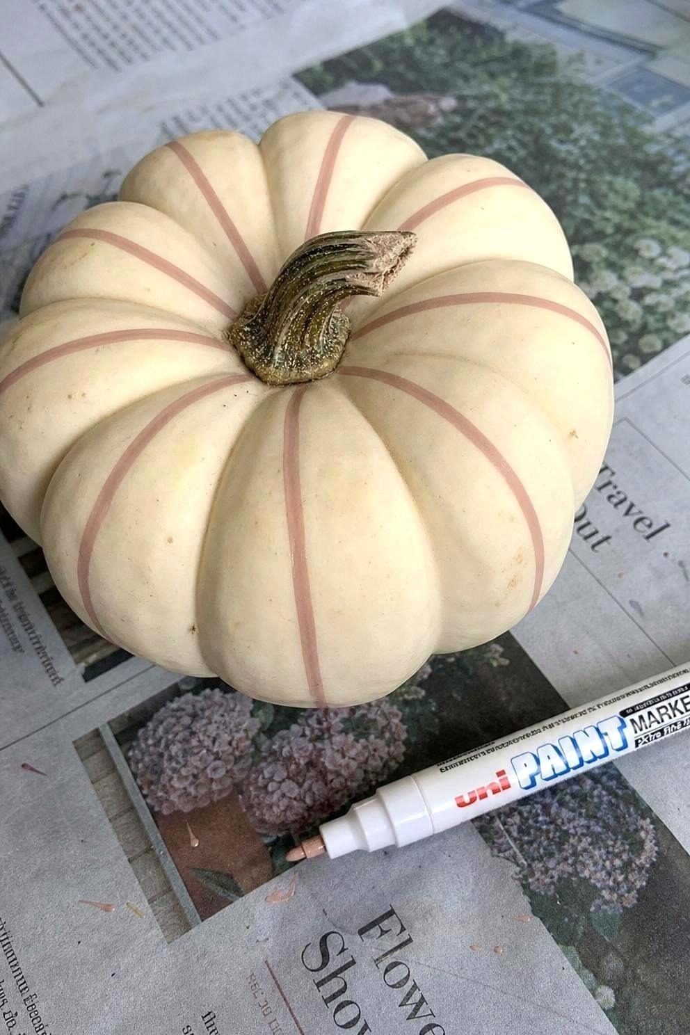 Close view of a cream pumpkin with simple paint-pen stripes sitting on newspaper next to one paint pen