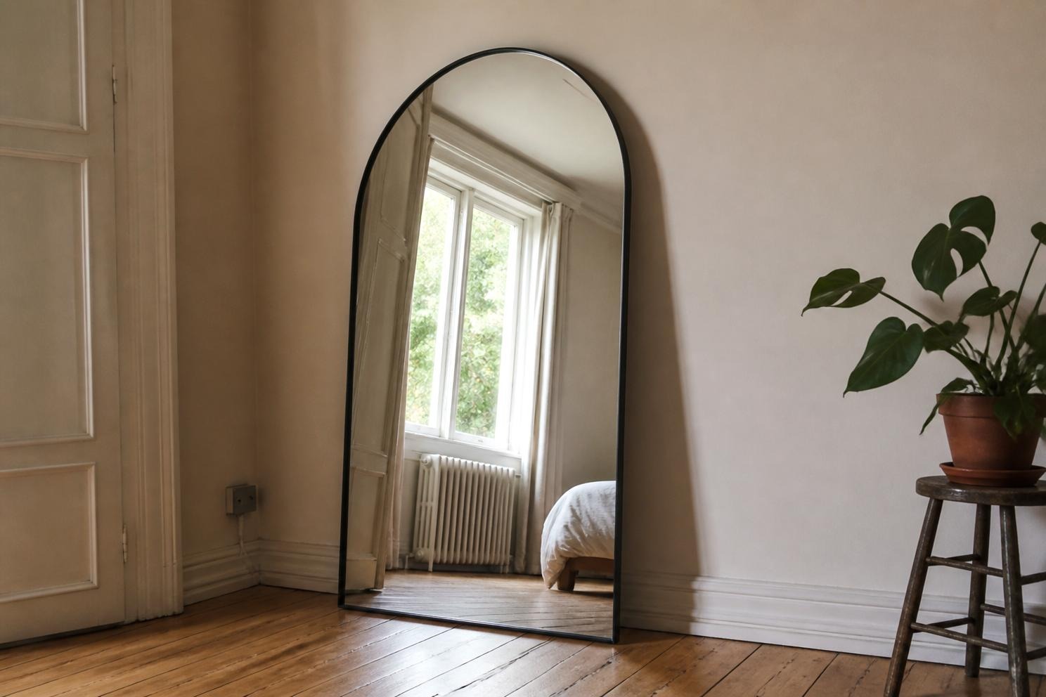 A tall arched mirror with a slim dark frame leaning against a wall facing a window