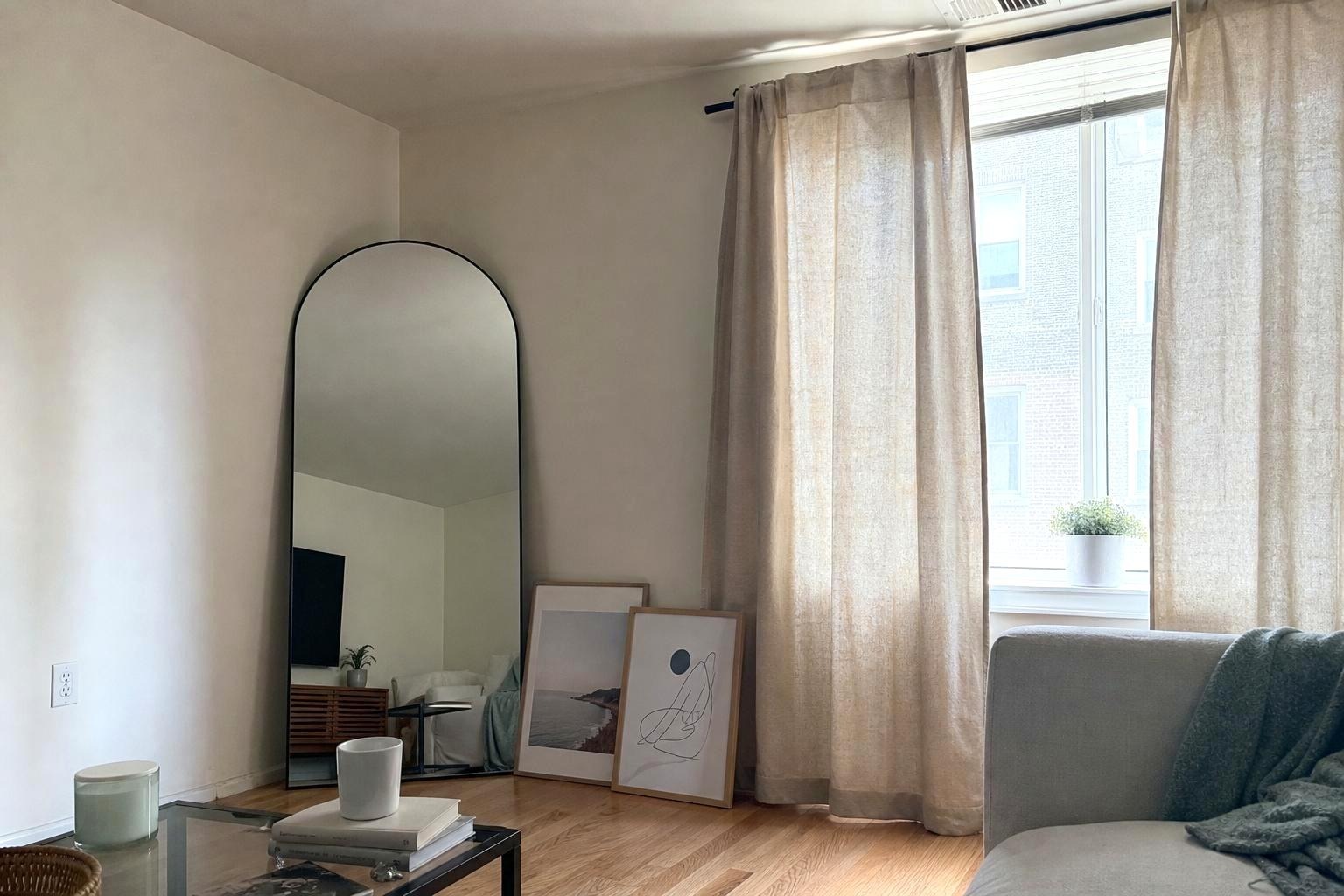A rented living room wall with a leaning framed print, a tension rod curtain, and a swapped brass switch plate cover