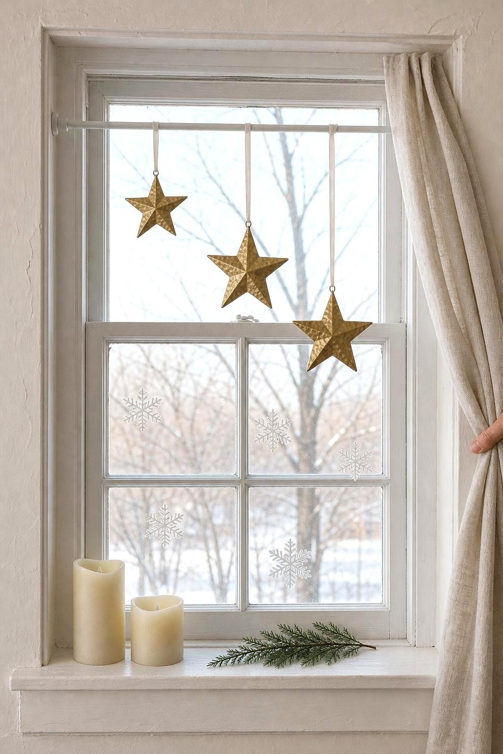 A rented window with a white tension rod wedged inside the reveal holding three brass stars on ribbon at staggered drops, snowflake clings on the lower panes