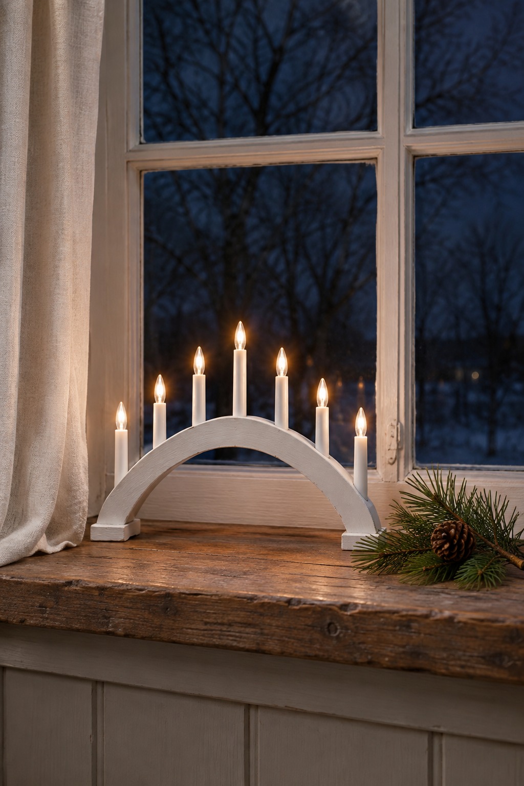 White wooden candle bridge with seven lit candle lights on a windowsill, a pine sprig beside it and a dark winter window behind