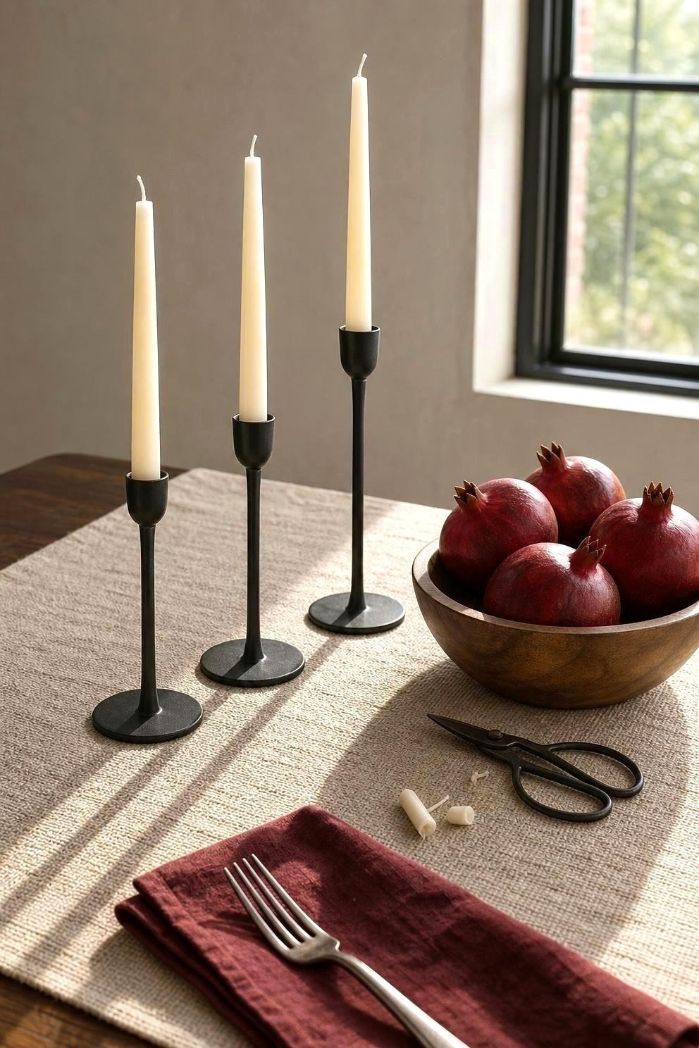 Three black metal taper candle holders of different heights on a linen runner, holding unlit ivory tapers, with a small bowl of pomegranates beside them