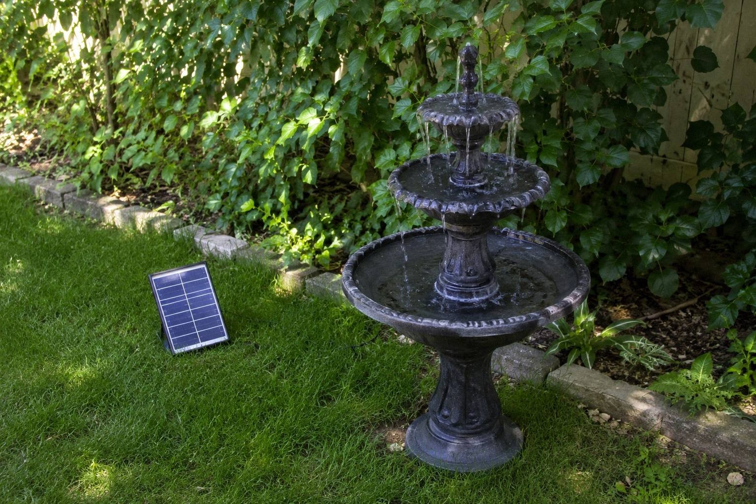 Three-tier solar fountain bird bath with moving water in a green backyard corner, small solar panel resting on the grass