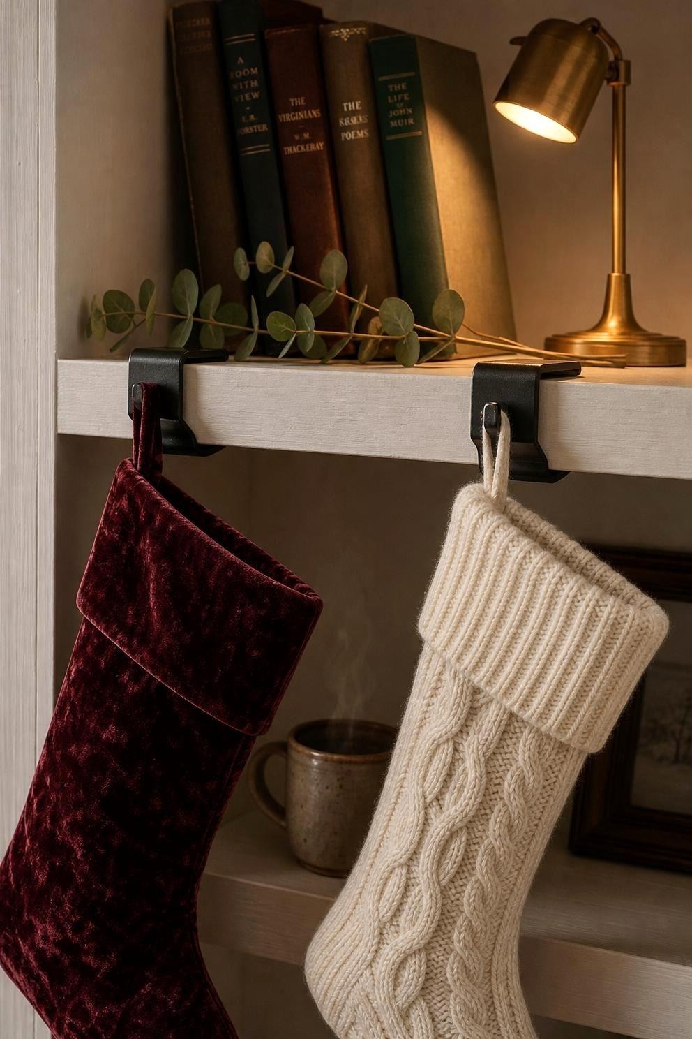 Two knitted stockings hanging from black clamp-style holders gripping the edge of a wooden bookshelf, with books and a small brass lamp behind